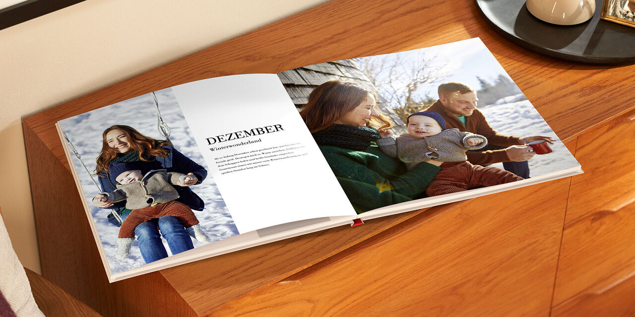 Le livre de photos ouvert est posé sur un meuble en bois. Sur la page de gauche, se trouvent une photo d’une femme et d’un enfant ainsi qu’un bloc de texte avec le titre « Décembre ». Sur la page de droite, la famille est représentée sur une photo pleine page.